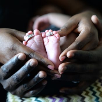 mother and father's hands around baby's feet with their wedding bands on the baby's toes.