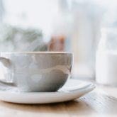 closeup photo of a steaming white ceramic coffee cup and glass jar of milk blurred in the background