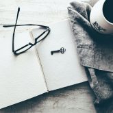 A pair of glasses and a small black key lie on top of a blank notebook. To the right is a grey cloth and a mug.