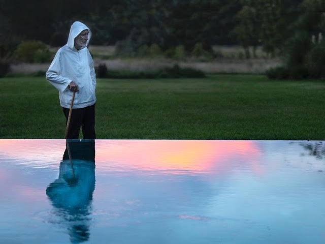Br. David Steindl-Rast wearing white shirt with a hood, holds a walking cane beside a reflecting pool