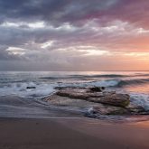 waves crashing against a sandy and rocky shoreline at sunset, with clouds of purple, pink and orange