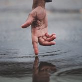 close up photo of a mans hand facing the camera with a finger outstretched creating a ripple effect in a pool of water